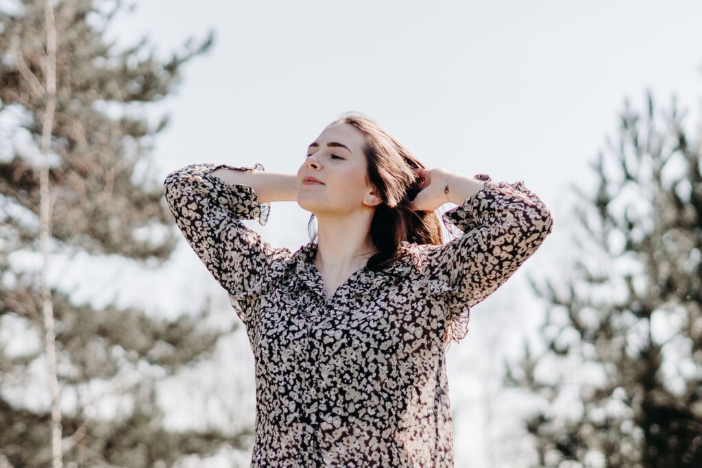 A woman stands outdoors, smiling with closed eyes, in a floral blouse, enjoying the serene environment.